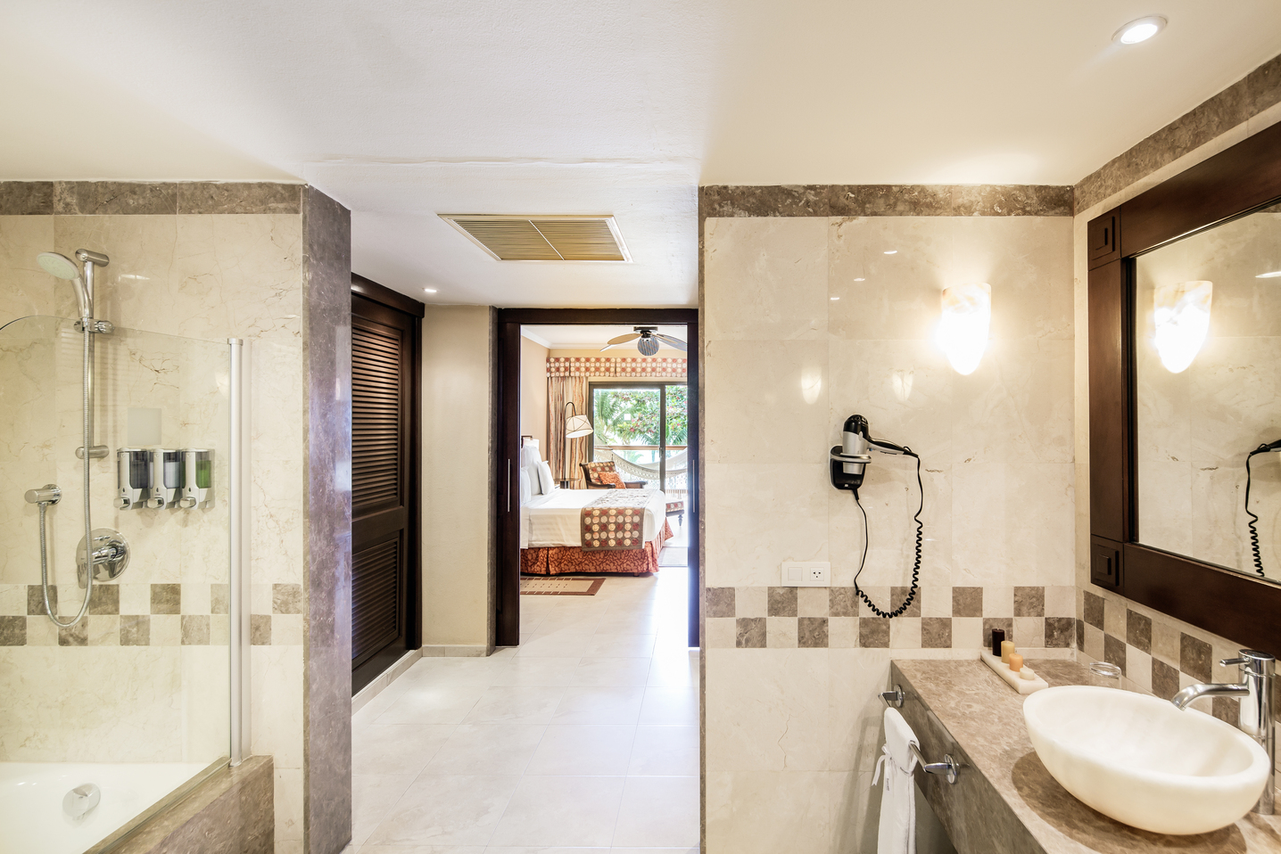 Bathroom with marble walls, glass shower stall, vessel sink, and doorway opening to bedroom with ocean view.