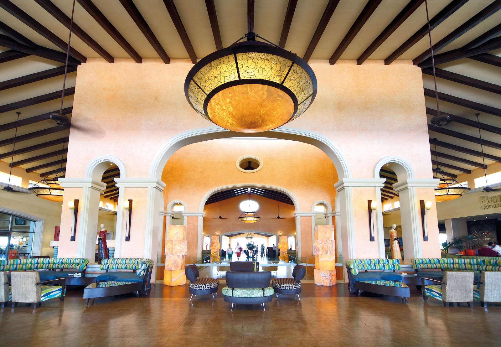 Grand lobby with striped wooden ceiling beams, arched doorways, orange accent walls, and colorful striped seating arranged around a central chandelier.