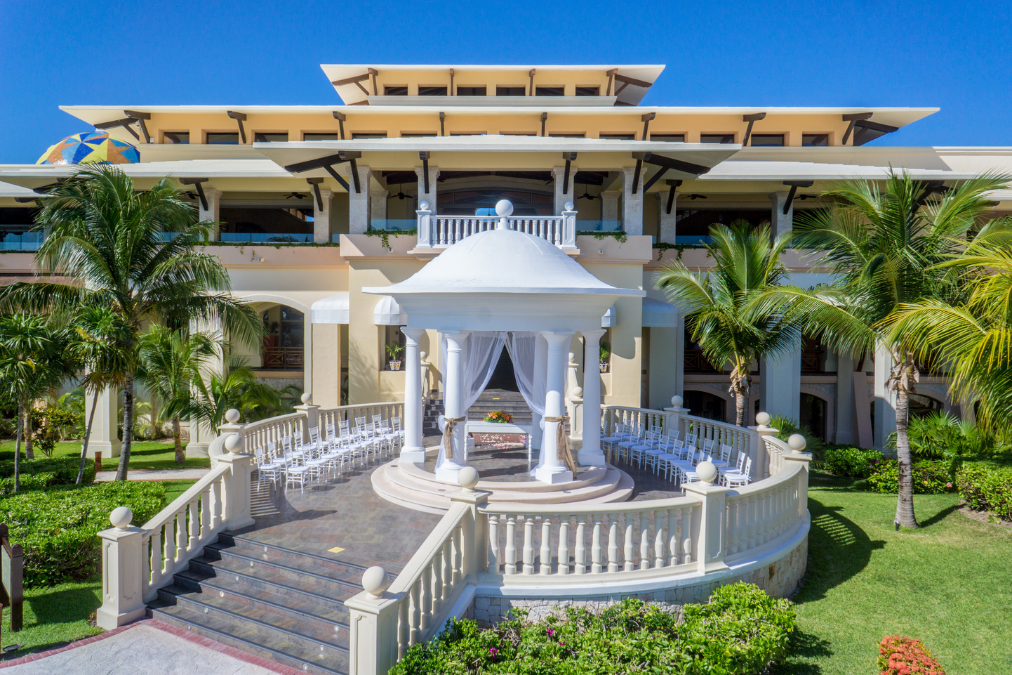 White domed gazebo with seated area surrounded by balustrades on manicured grounds, with multi-story resort building and palm trees in background.