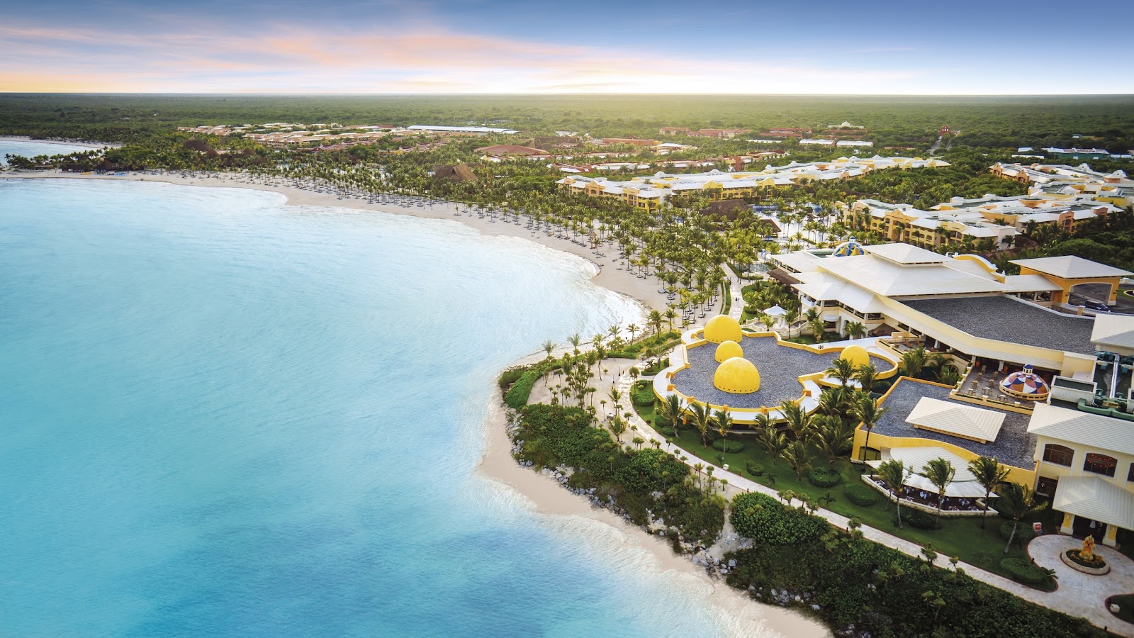 Aerial view of Barceló Maya Palace beachfront with turquoise water, white sand beach, yellow umbrellas, and palm-lined grounds extending into tropical forest.