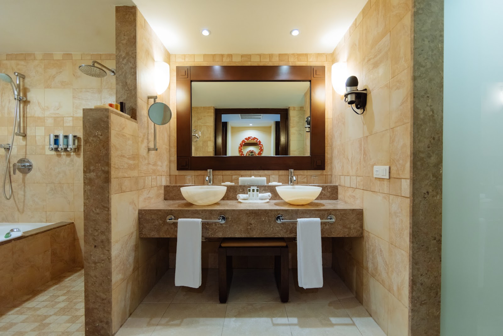 Spacious bathroom with dual vessel sinks, large mirror, travertine tile, shower area, and decorative wreath in the mirror frame.