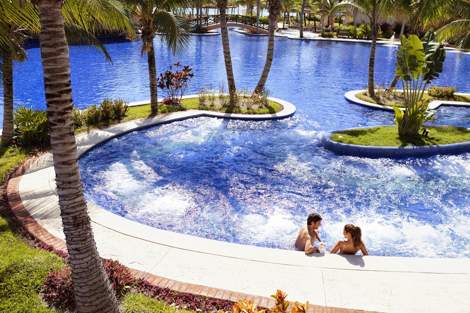 Two guests wade in a curved blue pool surrounded by palm trees and manicured landscaping at Barceló Maya Palace.