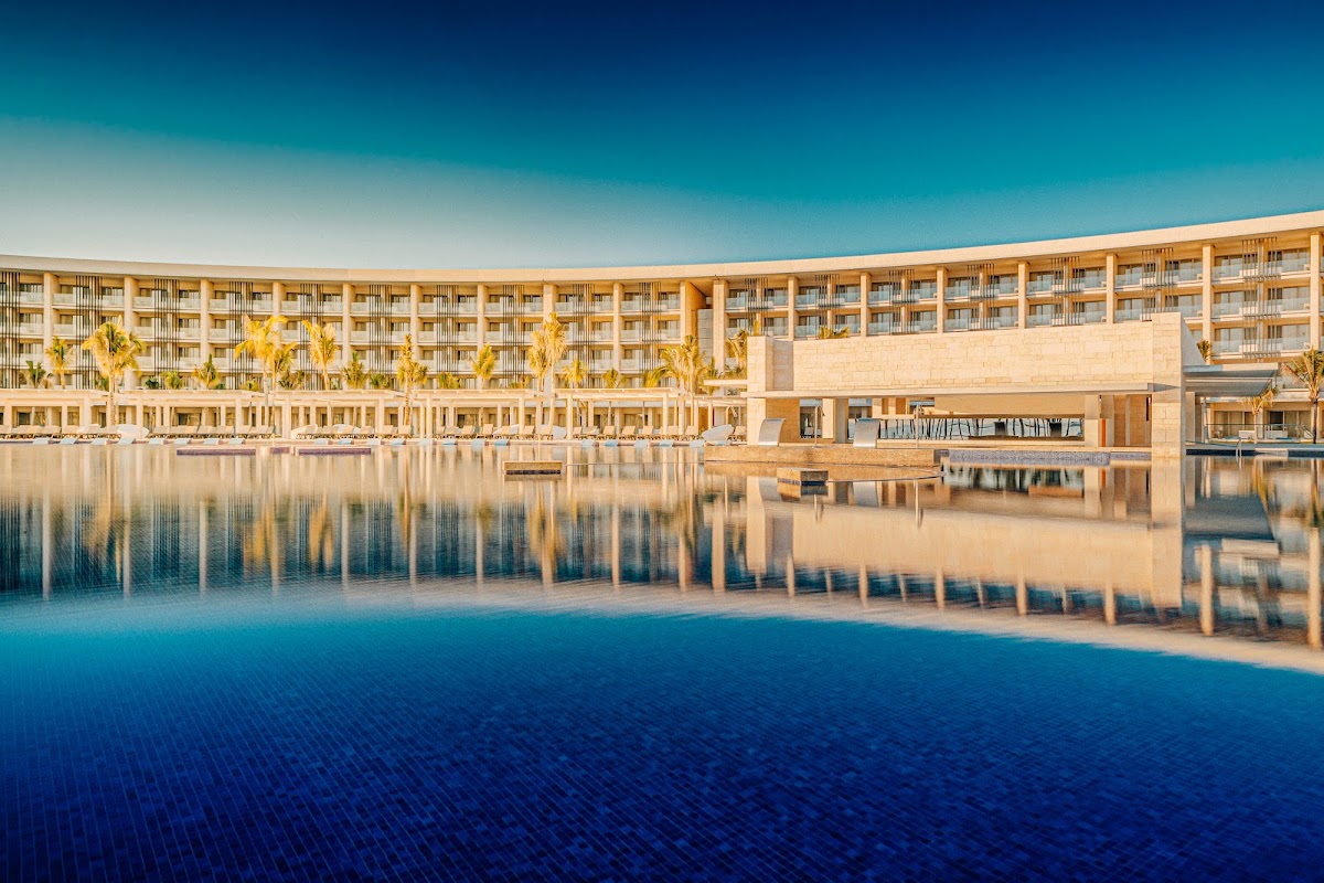 Large outdoor pool reflecting a modern beige resort building with palm trees under clear blue sky.