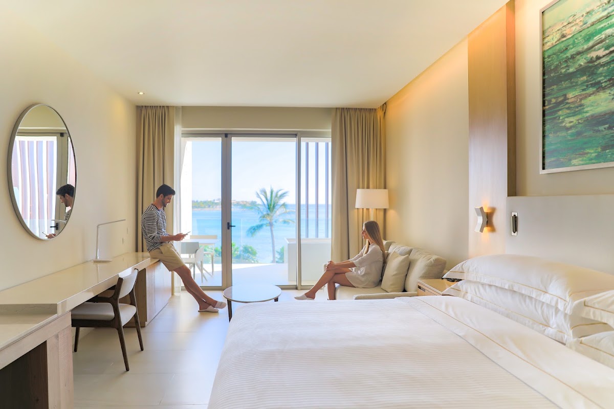 Two guests in a modern beige bedroom with ocean and palm tree views through sliding glass doors overlooking the beach.