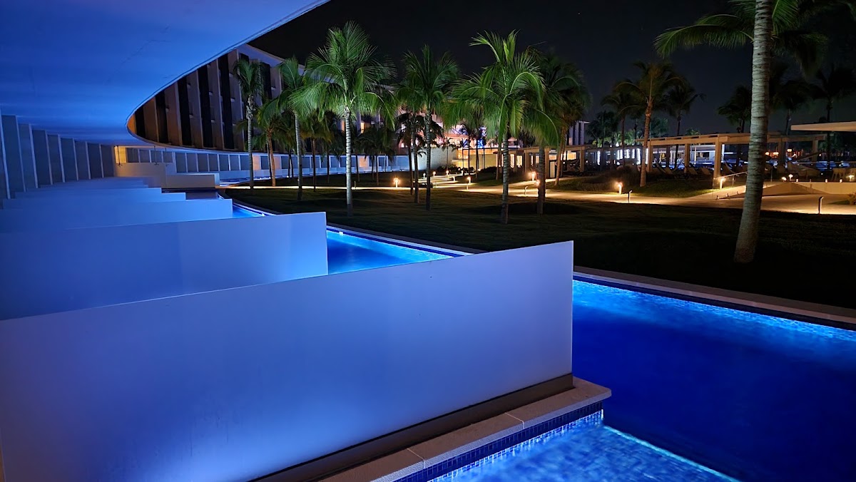 Illuminated pool with white walls at dusk, palm trees and lit buildings reflected in dark water beyond.