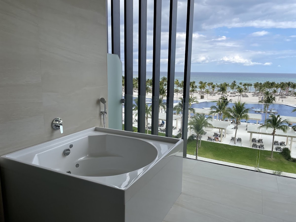 Corner soaking tub in a bathroom with floor-to-ceiling windows overlooking a pool, beach, and palm trees at Barceló Maya Riviera.