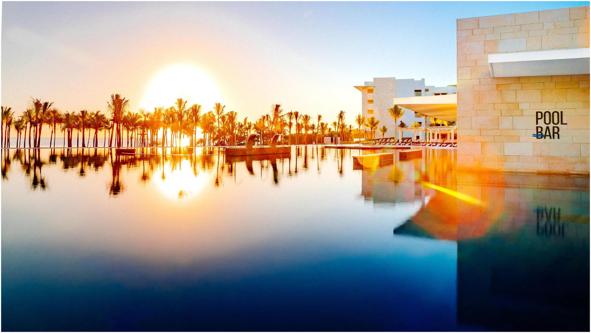 Resort pool bar at sunrise with palm trees reflected in still water and modern stone building at right.