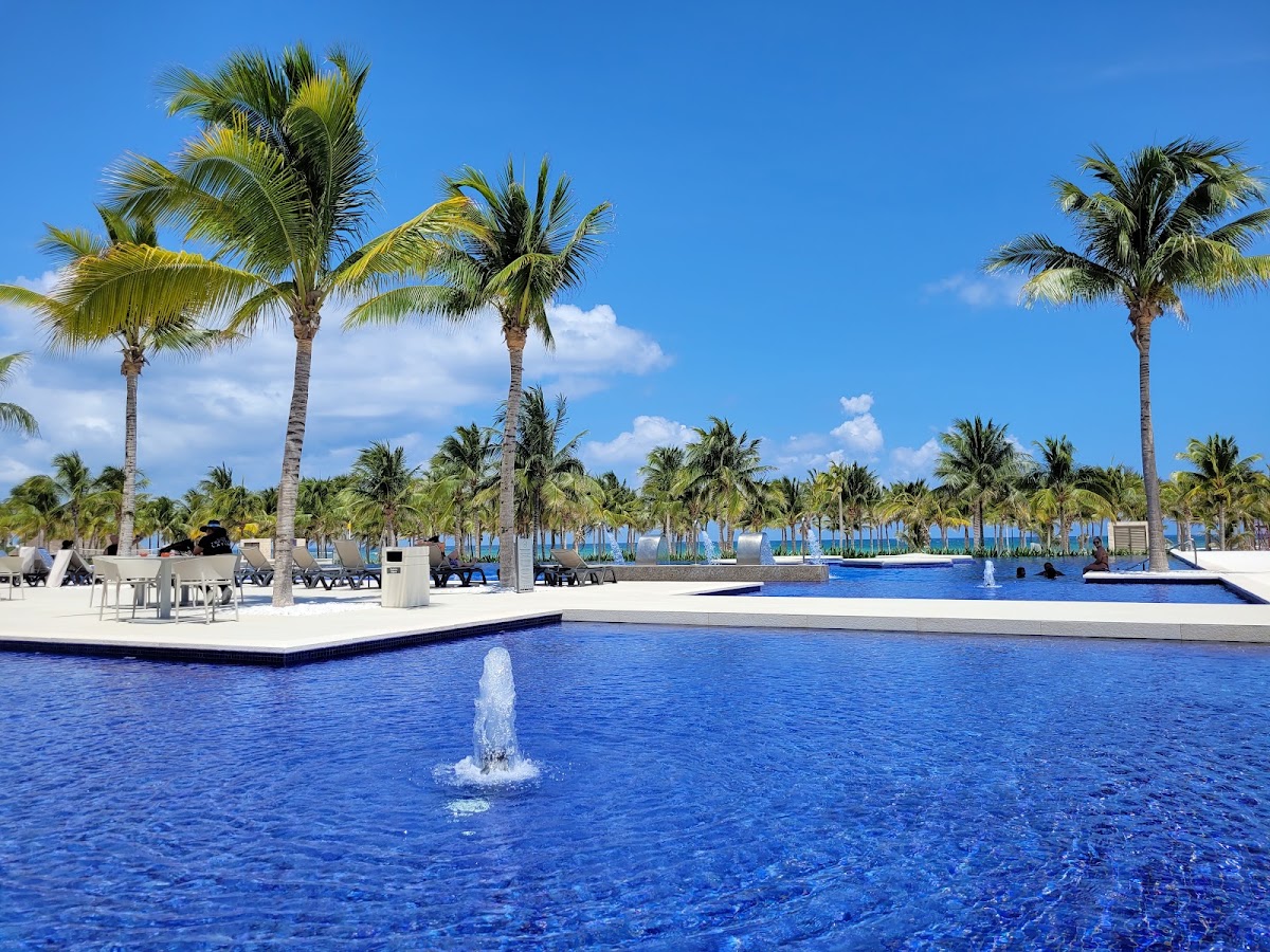 Resort pool with fountain and palm trees lining the deck, blue sky and white clouds above.