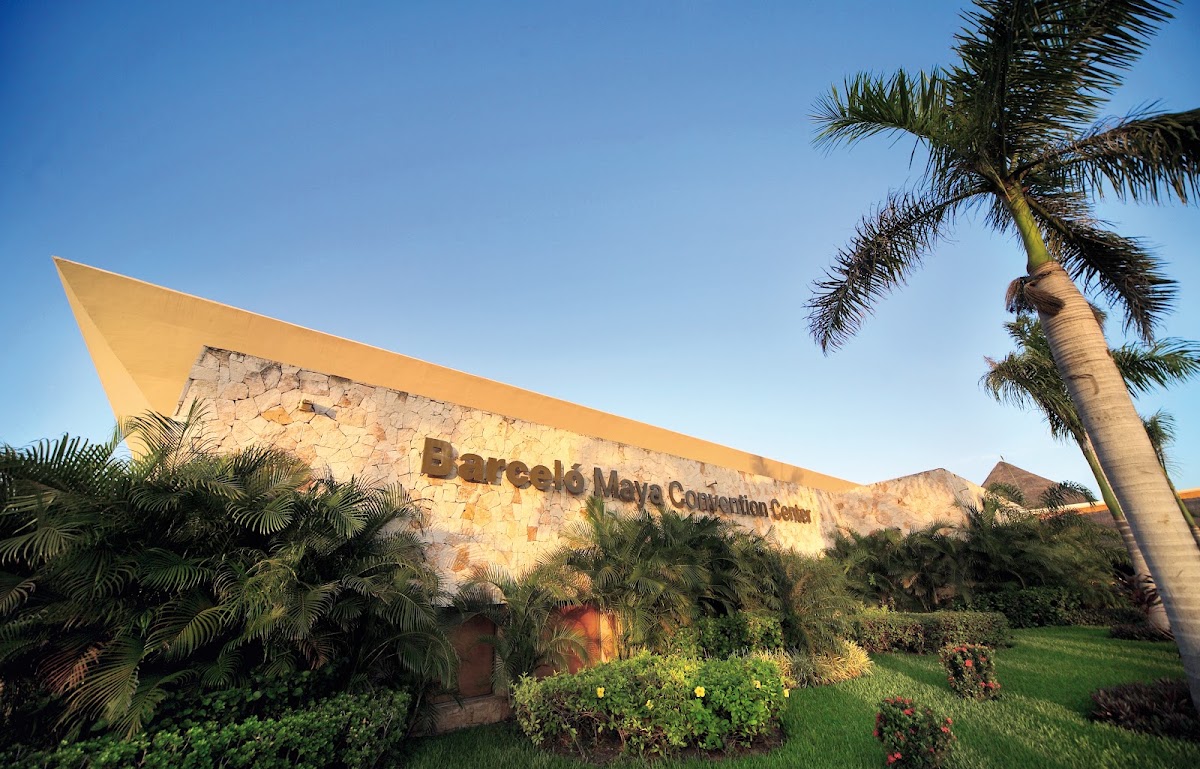 Barceló Maya Tropical Convention Center exterior with stone facade, yellow canopy, palm trees, and landscaped grounds under clear sky.