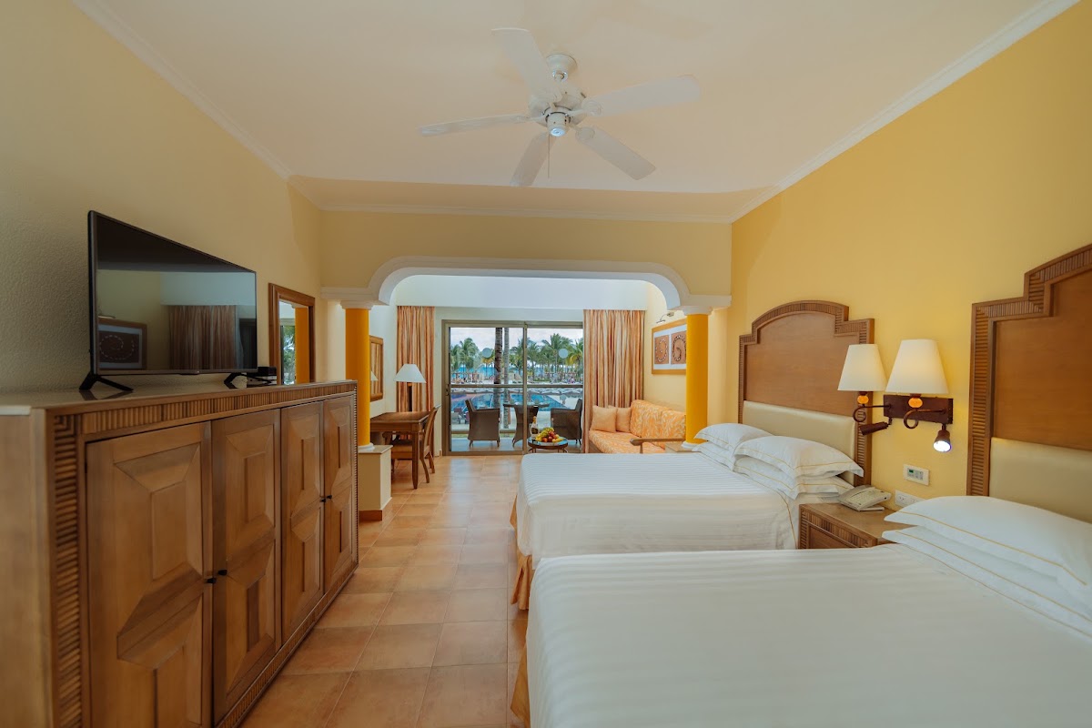 Resort room with two beds, wood furnishings, yellow walls, and balcony overlooking pool and palm trees at Barceló Maya Tropical.