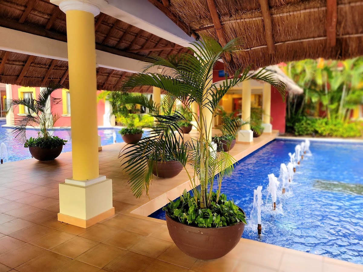 Covered walkway with yellow column and potted palms alongside a blue pool with fountains at Barceló Maya Tropical.