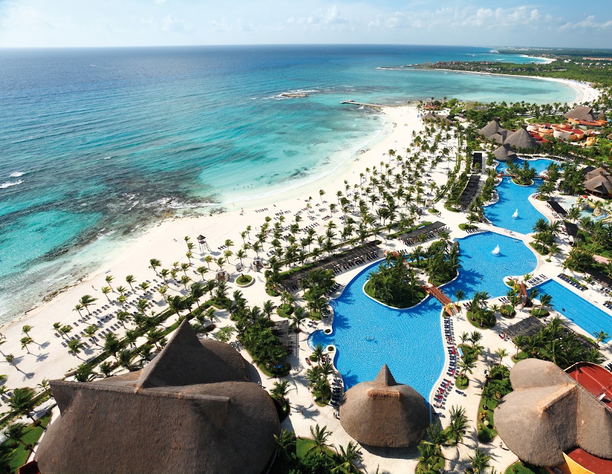 Aerial view of Barceló Maya Tropical's beachfront with turquoise pools, white sand beach, thatched-roof buildings, and clear Caribbean waters.