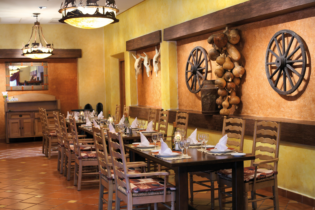 Rustic-themed dining room with yellow walls, wooden ladder-back chairs, wagon wheels, and dried gourds on terra cotta backdrop.