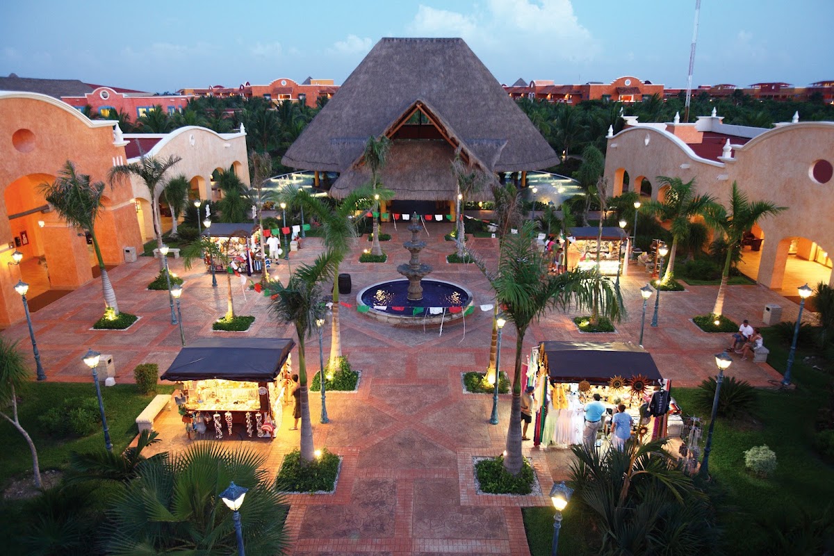 Overhead view of resort plaza at dusk with thatched-roof central structure, orange buildings, palm trees, fountain, and lit vendor stalls.