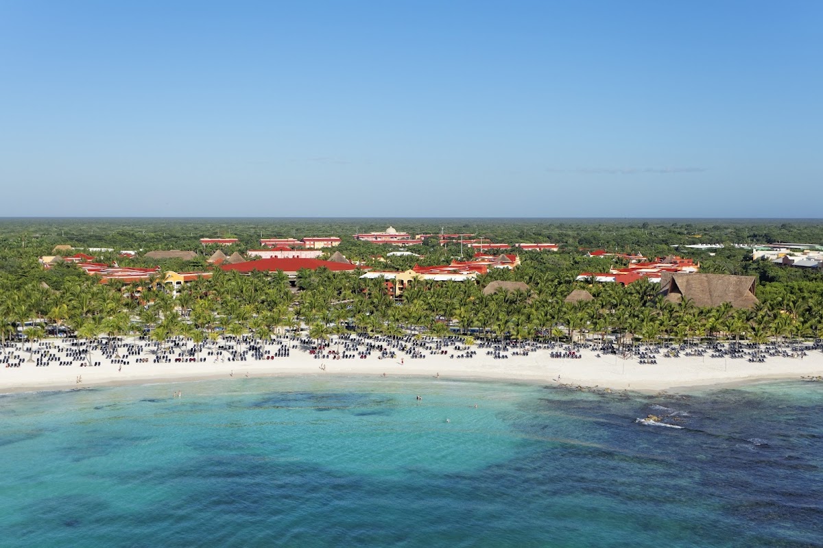 Barceló Maya Tropical beach resort with red-roofed buildings, palm trees, and white sand beach lined with blue umbrellas fronting turquoise water.