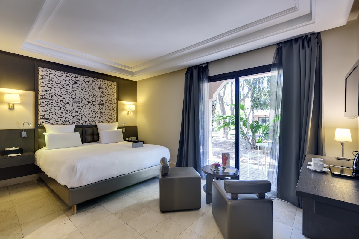 Modern resort bedroom with white bedding, patterned headboard, dark furnishings, and glass doors opening to a courtyard with palm trees.