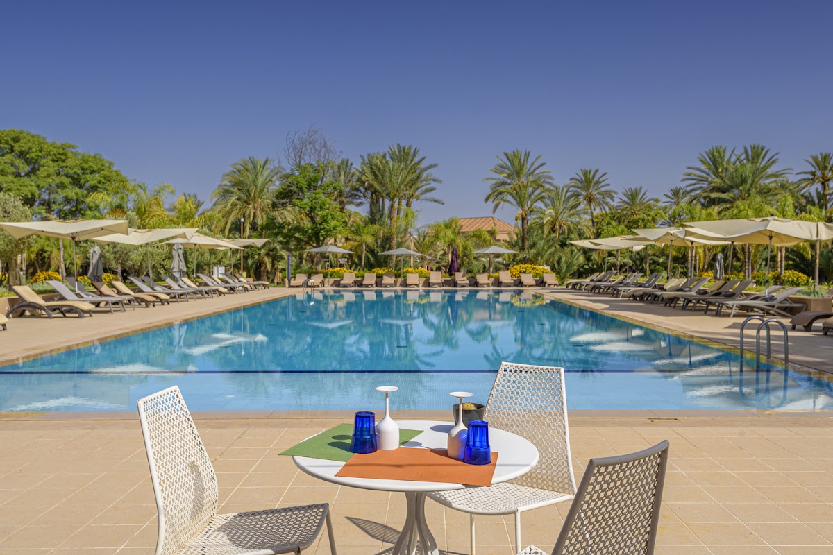 Large resort pool with loungers and cream umbrellas, surrounded by palm trees and clear blue sky at Barceló Palmeraie Oasis Resort.