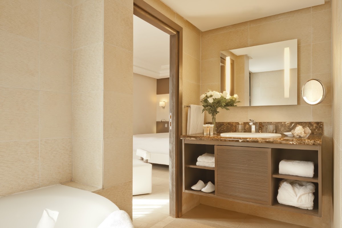 Bathroom with marble countertop, illuminated mirror, white flowers in a vase, and shelves stocked with rolled towels and toiletries.