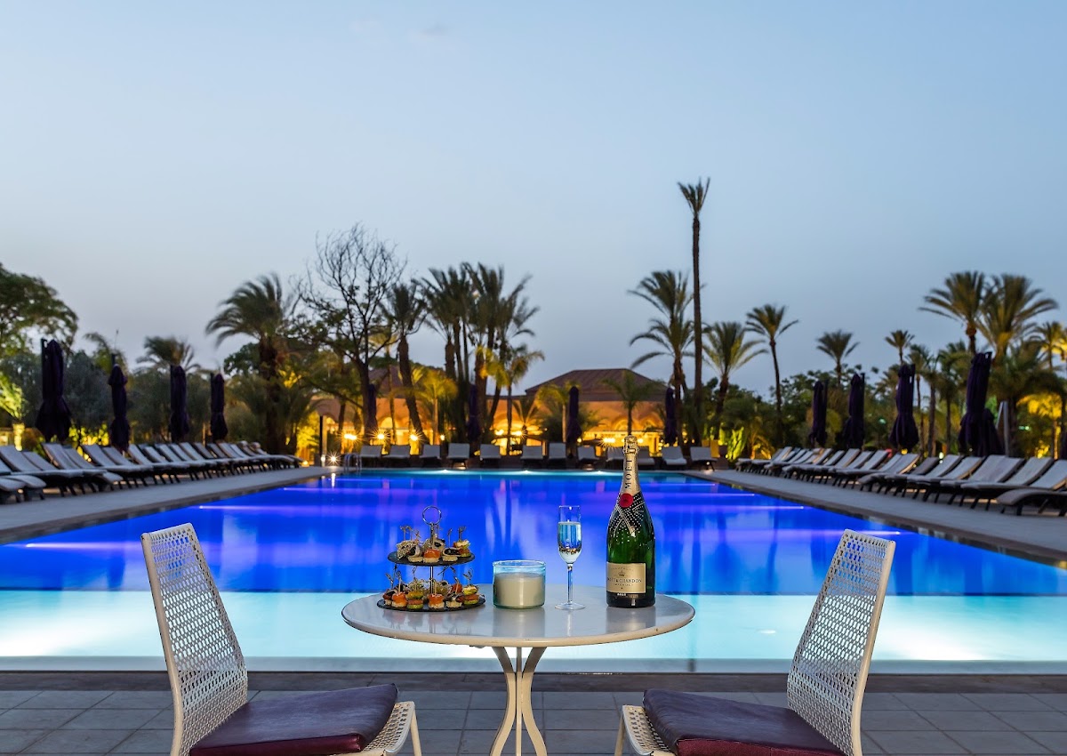 Poolside table set with champagne and appetizers at dusk, overlooking the illuminated blue pool surrounded by palms and lounge chairs at Barceló Palmeraie Oasis Resort.