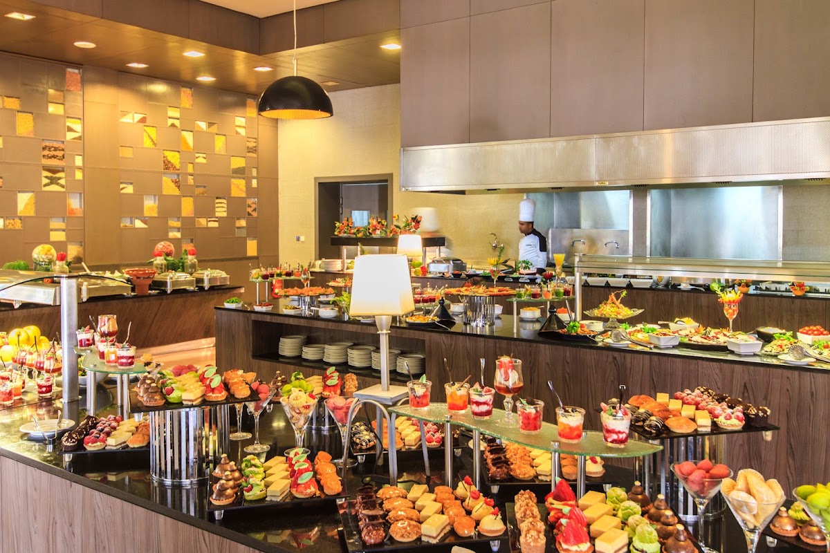 Chef prepares food behind buffet stations laden with appetizers, desserts, and tropical fruits in a modern dining hall with warm lighting.
