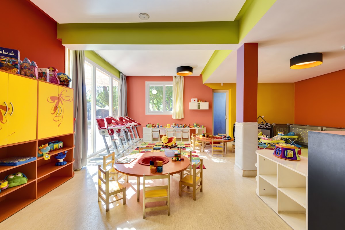 Colorful kids club room with yellow and orange storage units, round red table with chairs, toys, and bright geometric ceiling at Barceló Palmeraie Oasis Resort.