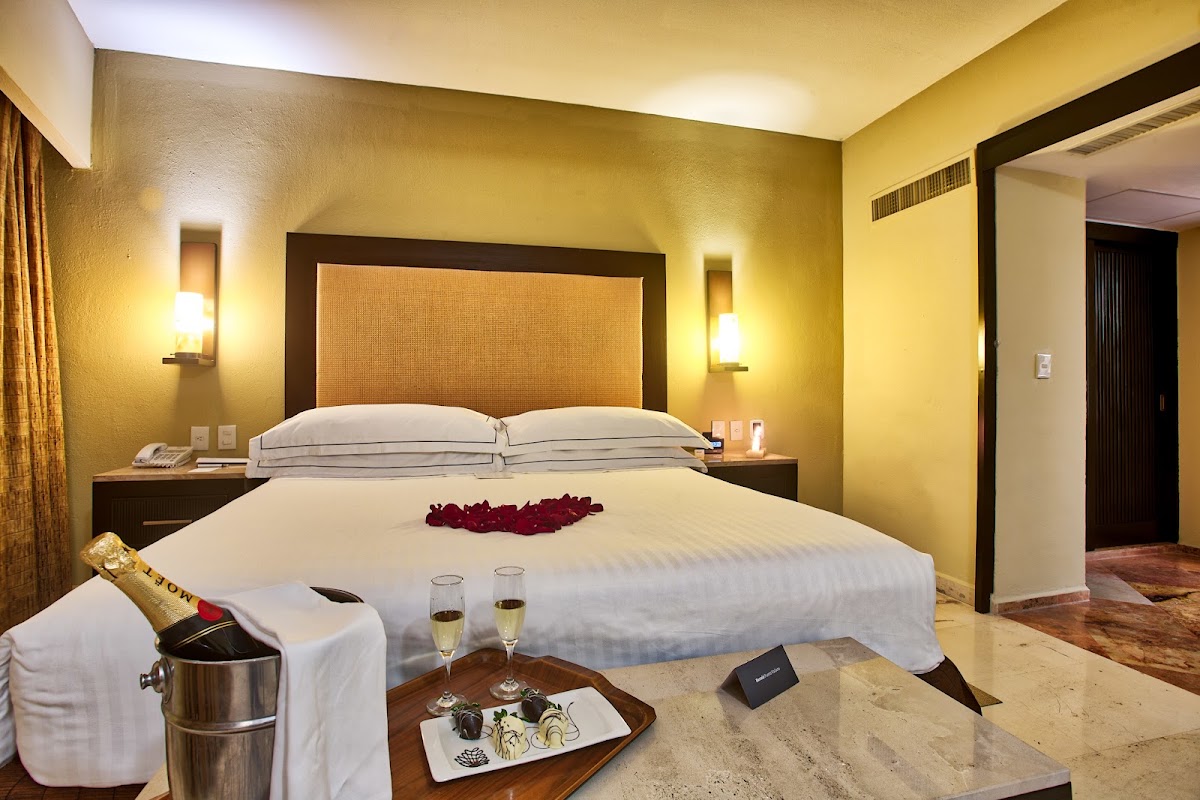 Resort suite with yellow walls, king bed with rose petals, champagne bucket, and two glasses on a table.