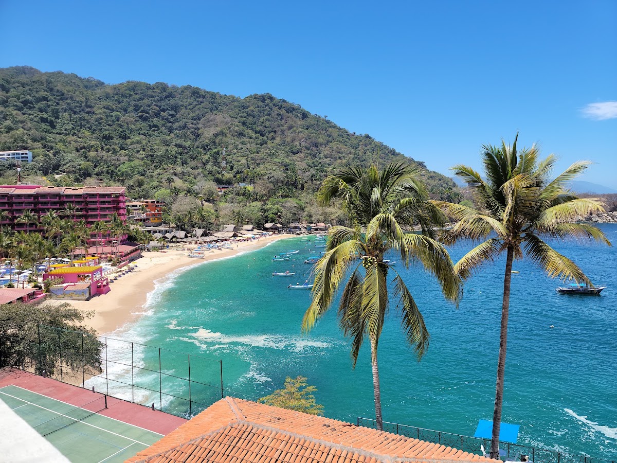 Pink resort building overlooks a sandy beach with turquoise water, palm trees, and forested hillside under clear blue sky.