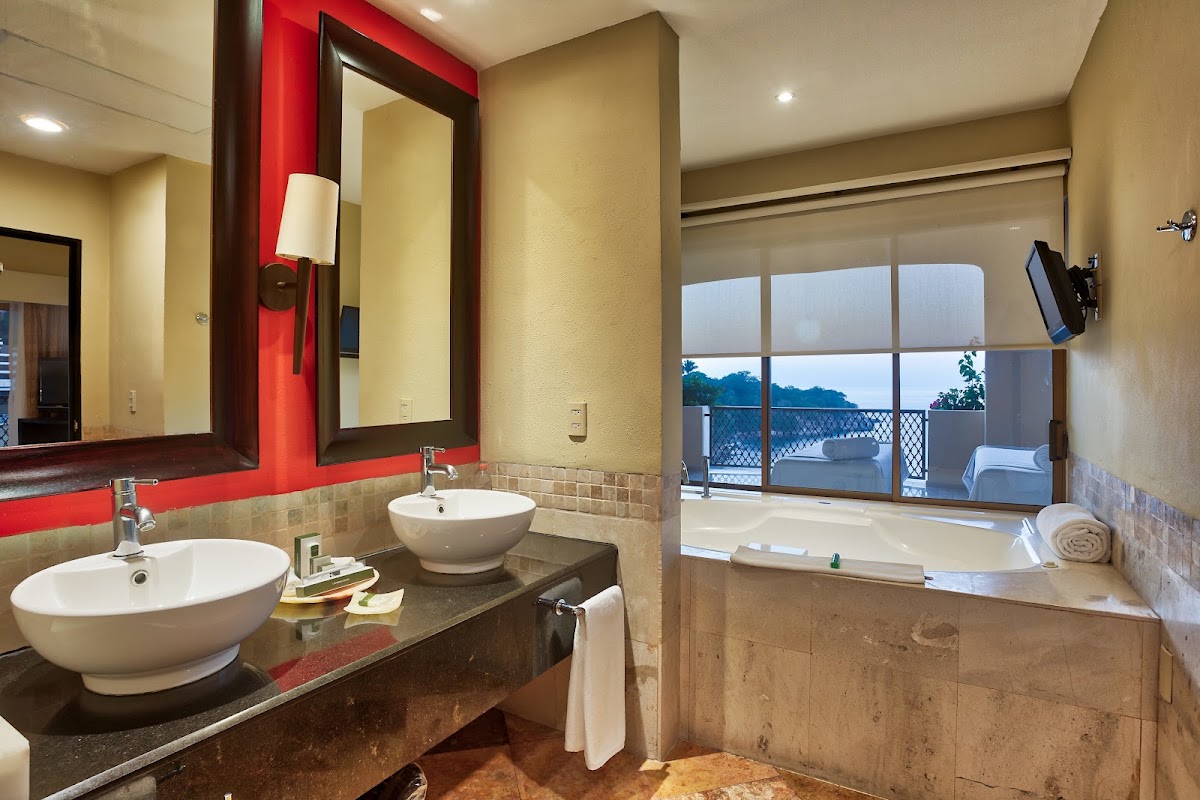 Resort bathroom with dual vessel sinks, red accent wall, and soaking tub overlooking ocean and balcony through glass doors.