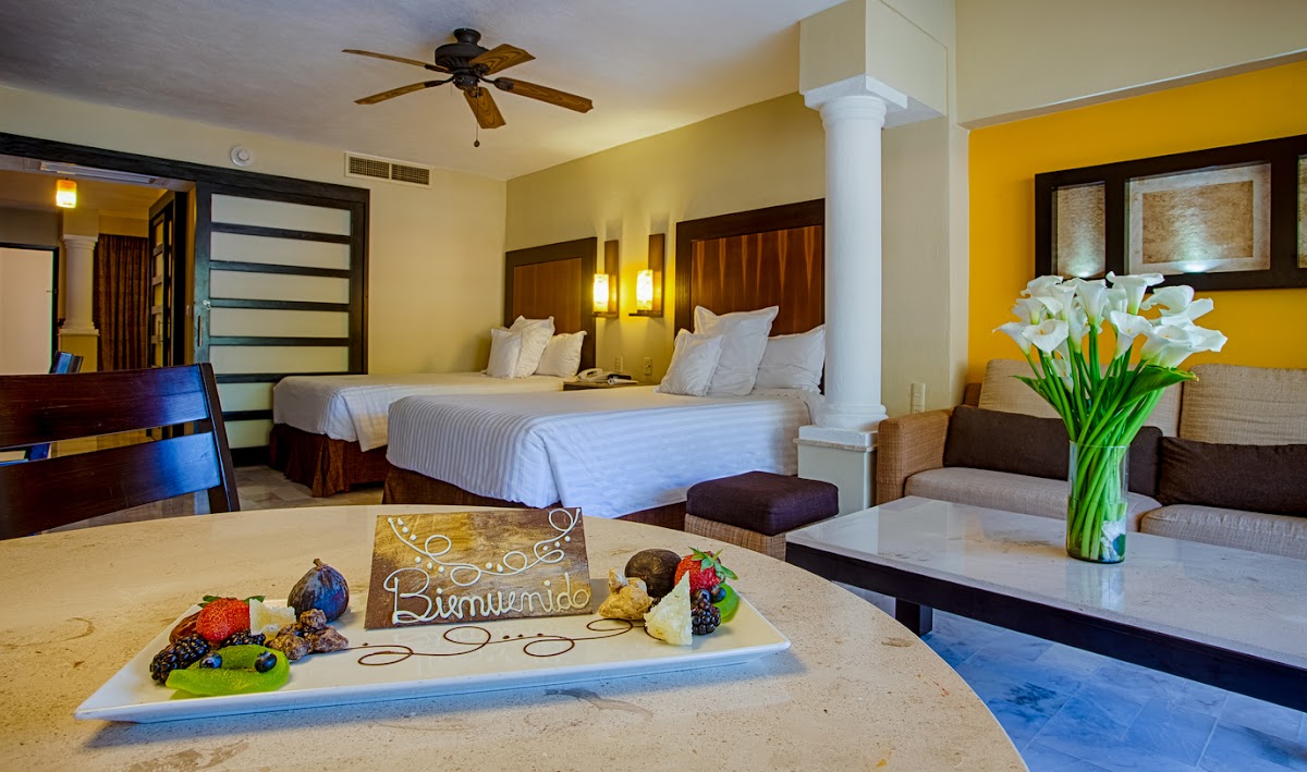 Resort guestroom with two queen beds, yellow walls, welcome fruit platter on counter, and sitting area with white flowers.