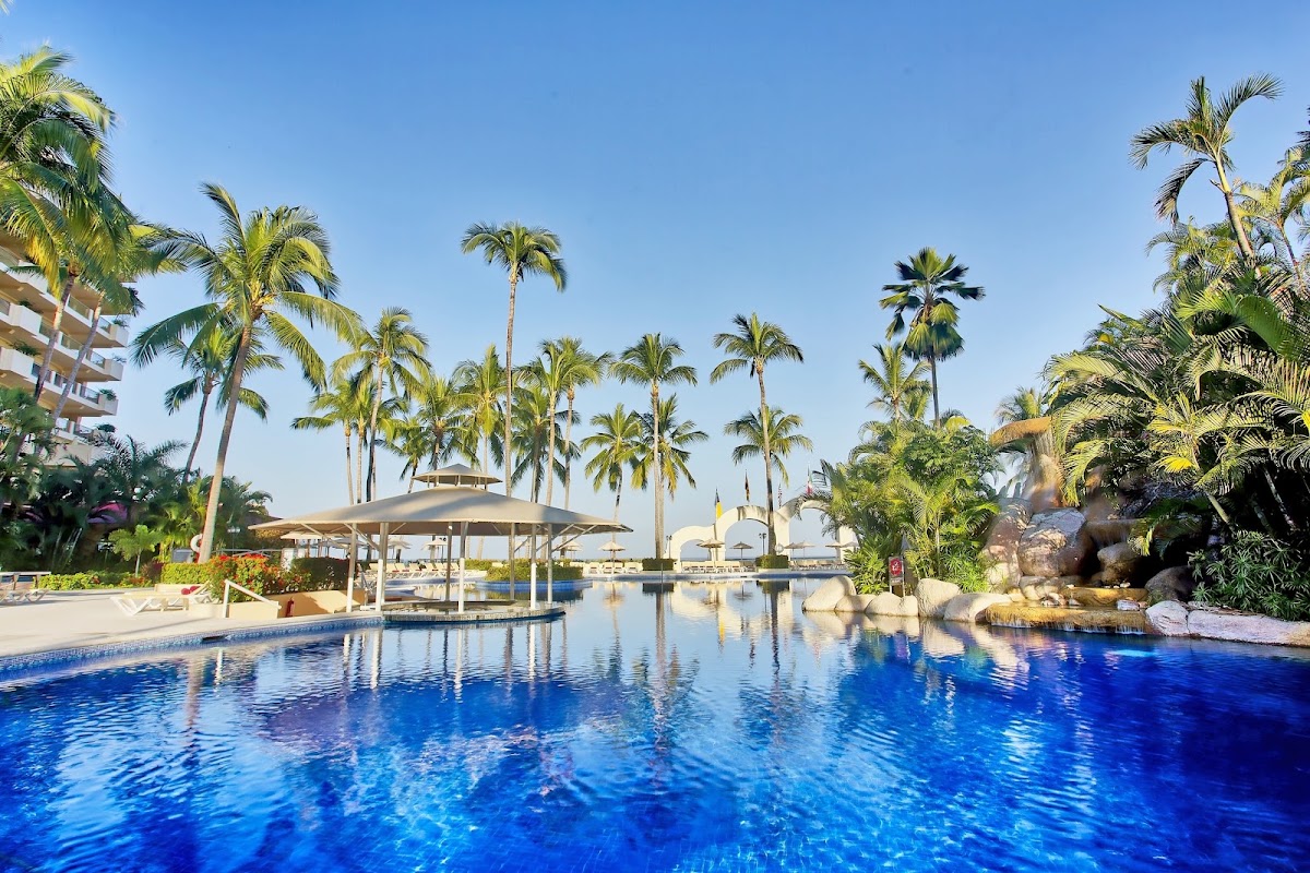 Blue pool with swim-up bar and tan umbrella surrounded by tall palm trees and resort buildings under clear sky.