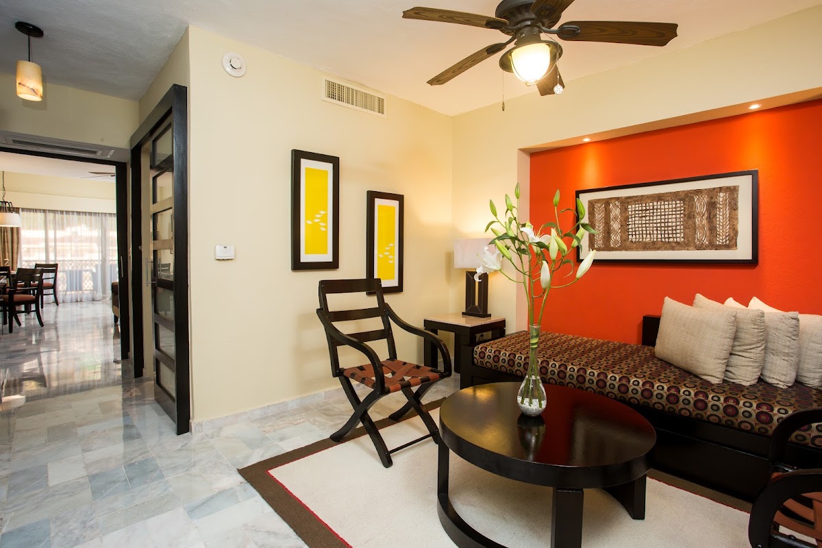 Resort suite with bright orange accent wall, black furniture, yellow framed artwork, and fresh flowers on dark table.