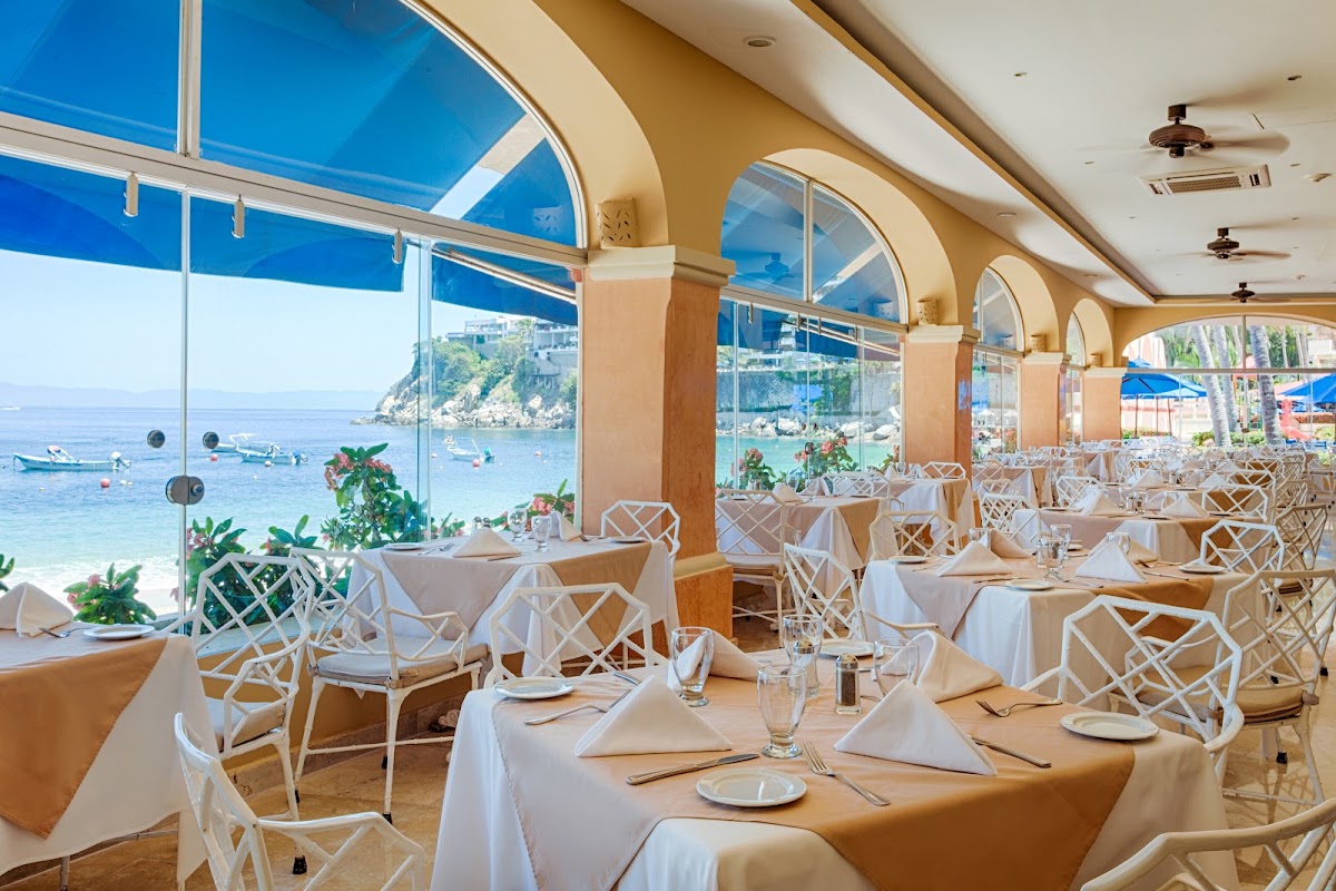 Dining room with arched windows overlooking a turquoise bay, white lattice chairs, tan tablecloths, and boats anchored in the water below.