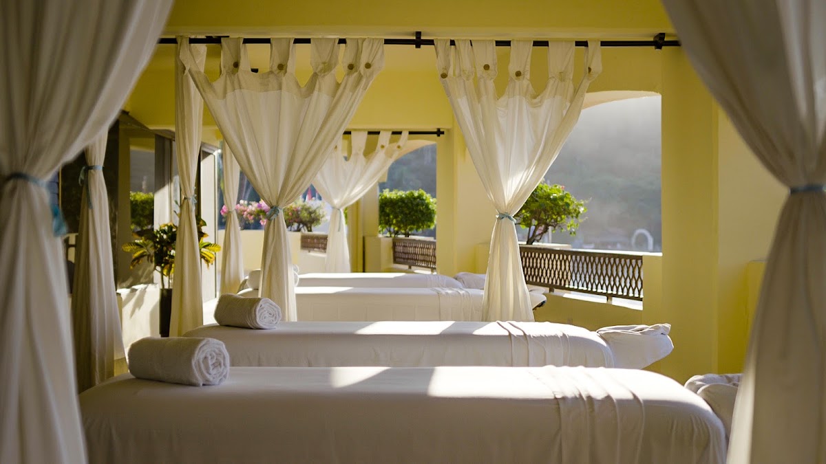 Spa treatment room with white canopy beds draped in sheer curtains, rolled towels, and mountain views visible through open windows at Barceló Puerto Vallarta.