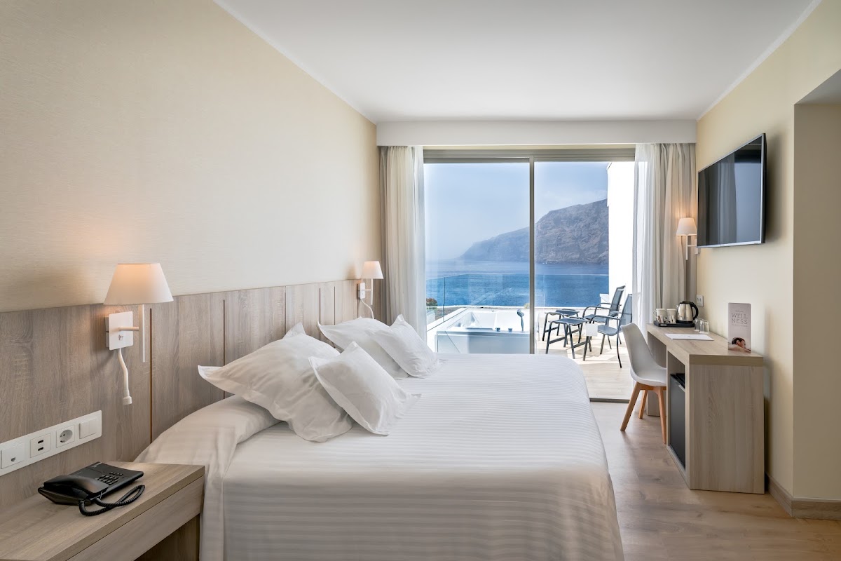 Hotel room with king bed, desk, and flat-screen TV overlooking a balcony with sea and rocky cliffs beyond.