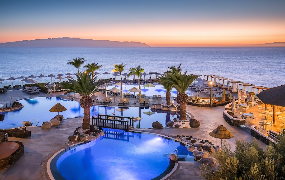 Illuminated pools and palm trees at Barceló Santiago overlook a beach with umbrellas and the sea at dusk.