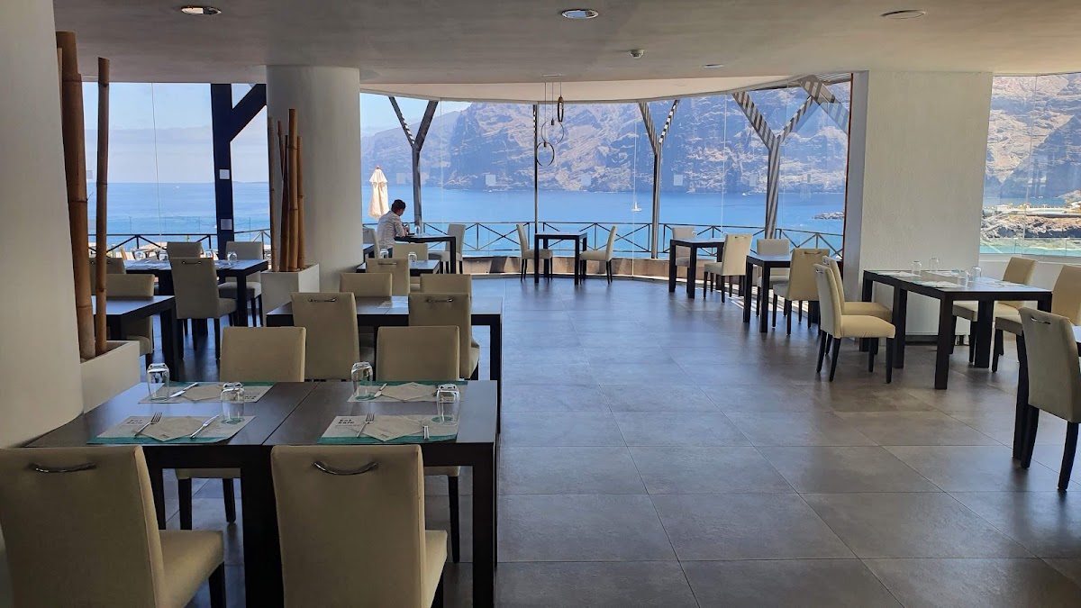 Dining room with cream chairs and dark tables overlooking a Mediterranean coastline and mountains through floor-to-ceiling windows.