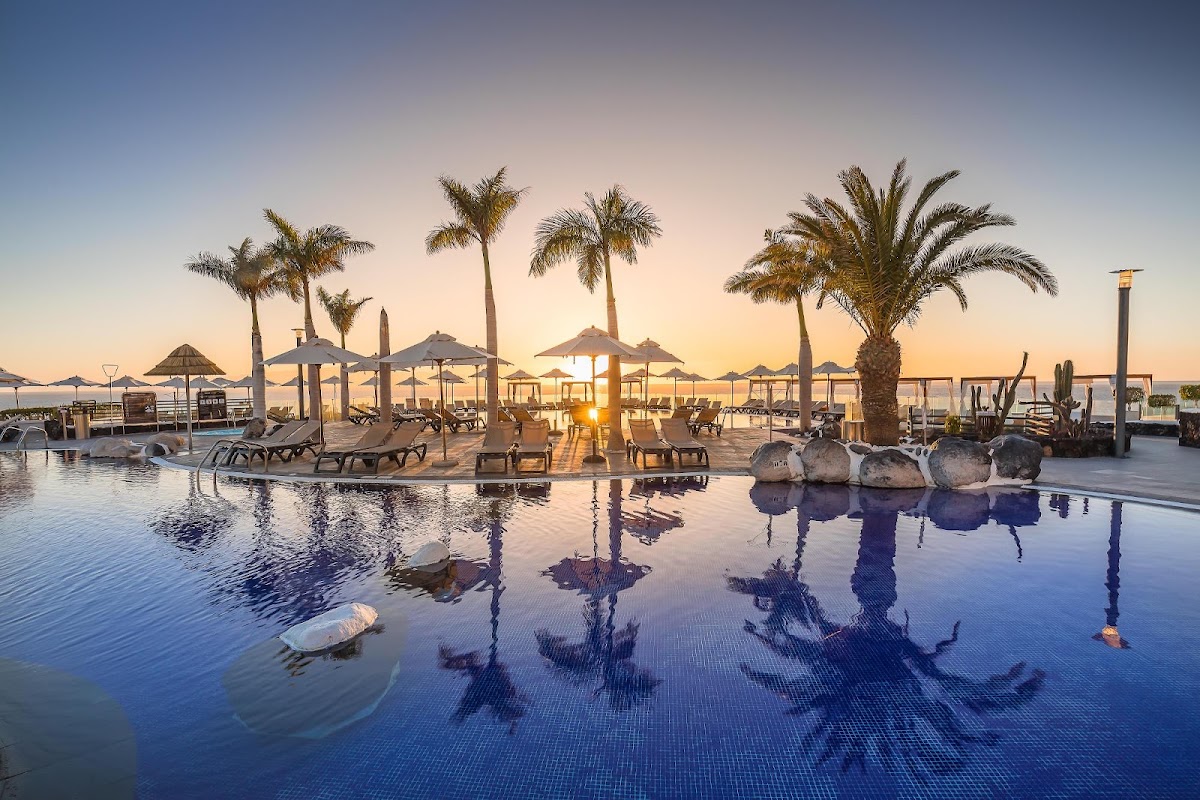 Pool area at dawn with loungers, thatched umbrellas, and palm trees reflected in still water at Barceló Santiago.