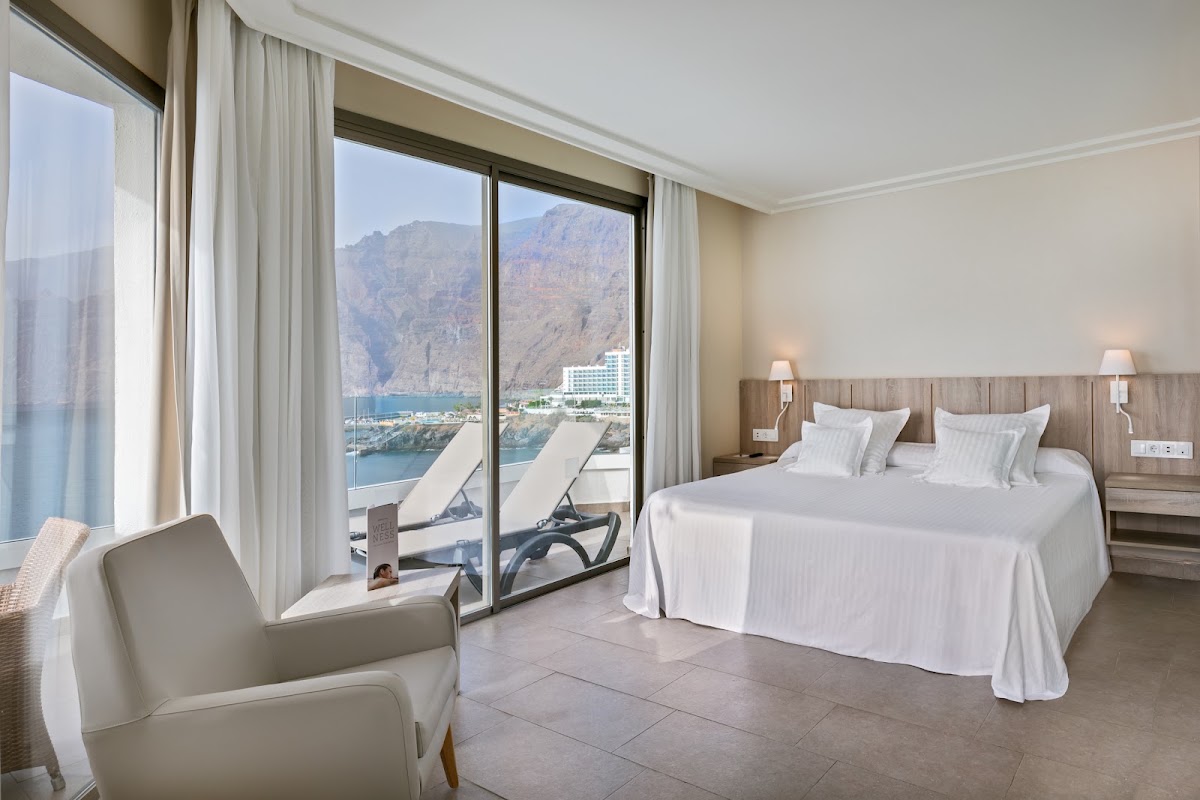 Resort bedroom with white bedding, cream armchair, and floor-to-ceiling windows overlooking a Mediterranean bay with mountains and a resort building.