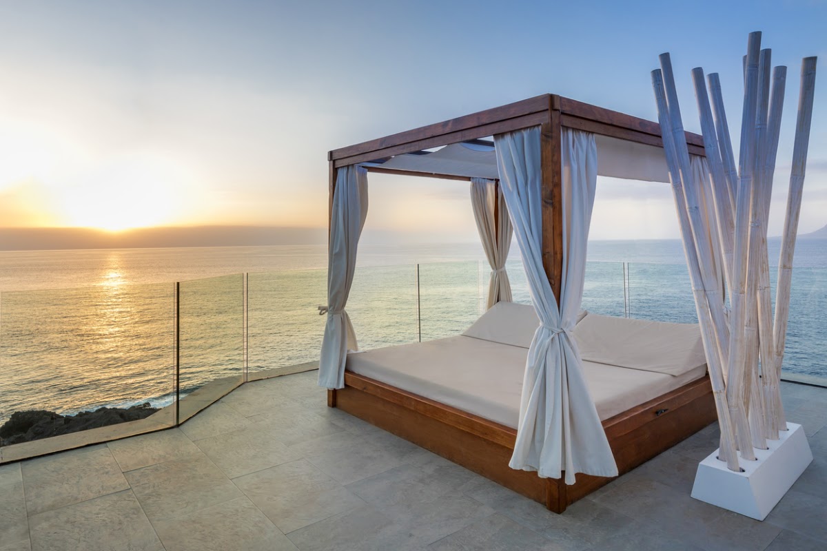 Wooden canopy bed with white curtains on a beachfront terrace overlooking the ocean at sunset.