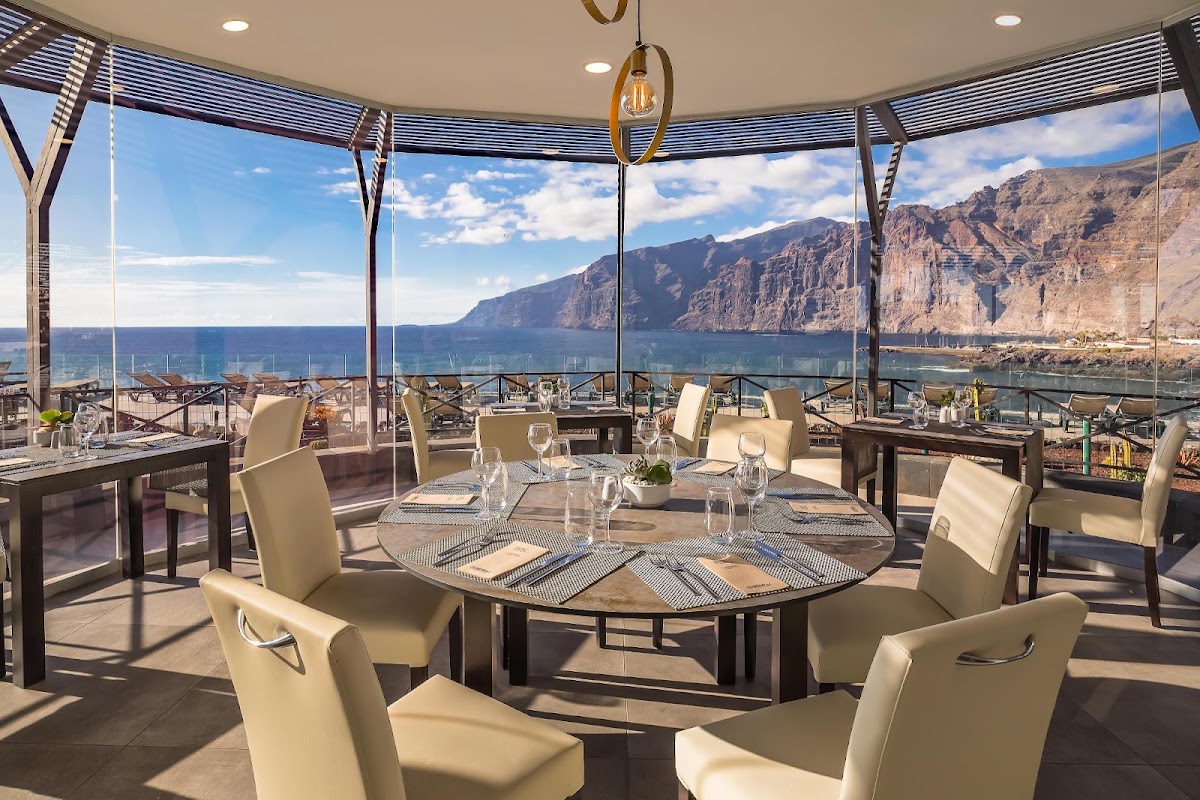 Dining room with round tables, cream chairs, and floor-to-ceiling windows overlooking rocky cliffs and blue ocean at Barceló Santiago.