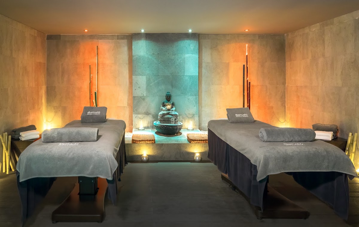 Spa treatment room with two massage beds flanking a central turquoise Buddha statue, warm orange-lit walls, and soft lighting throughout.
