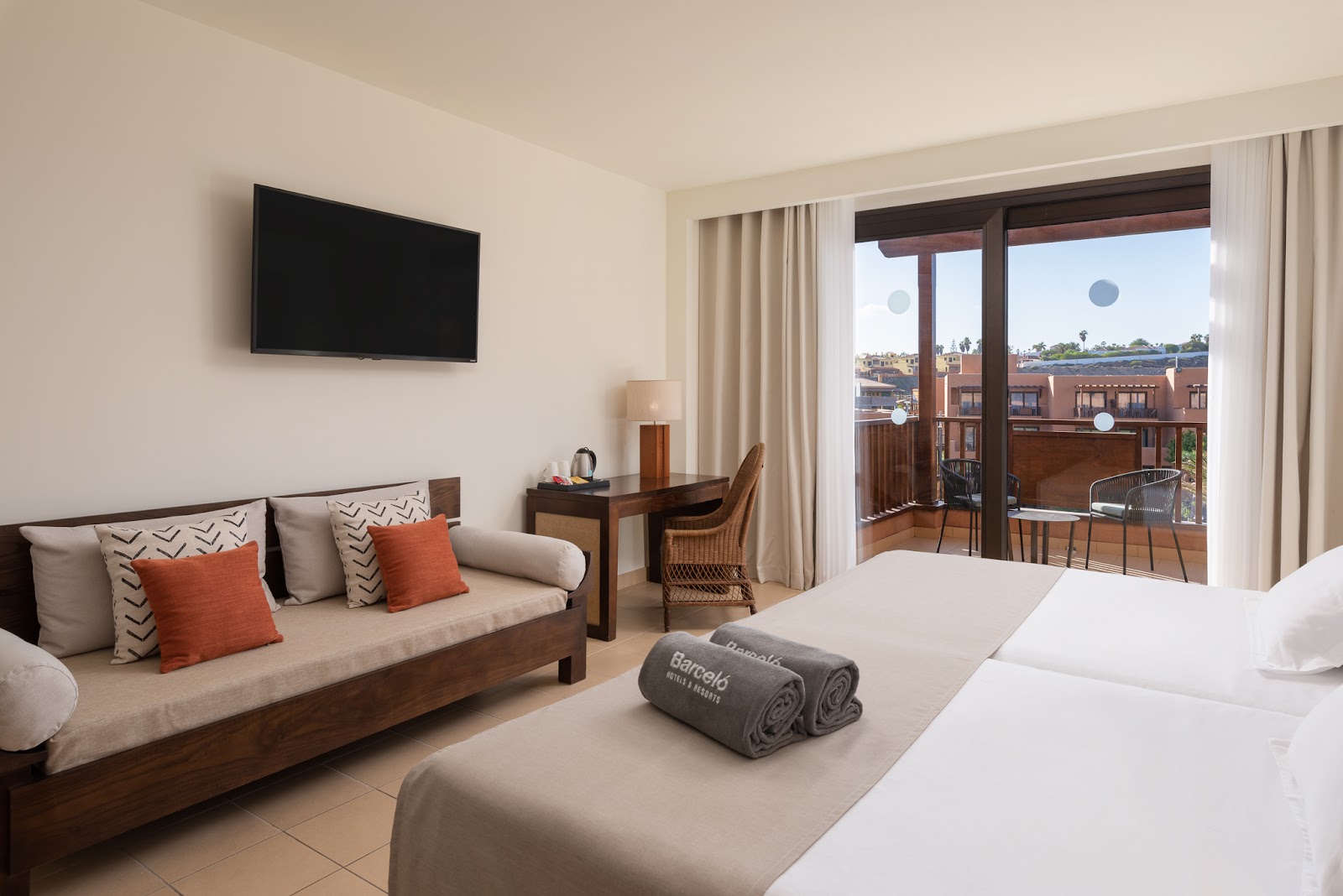 Resort guest room with bed, sofa with orange pillows, wall-mounted TV, and balcony overlooking Tenerife landscape.