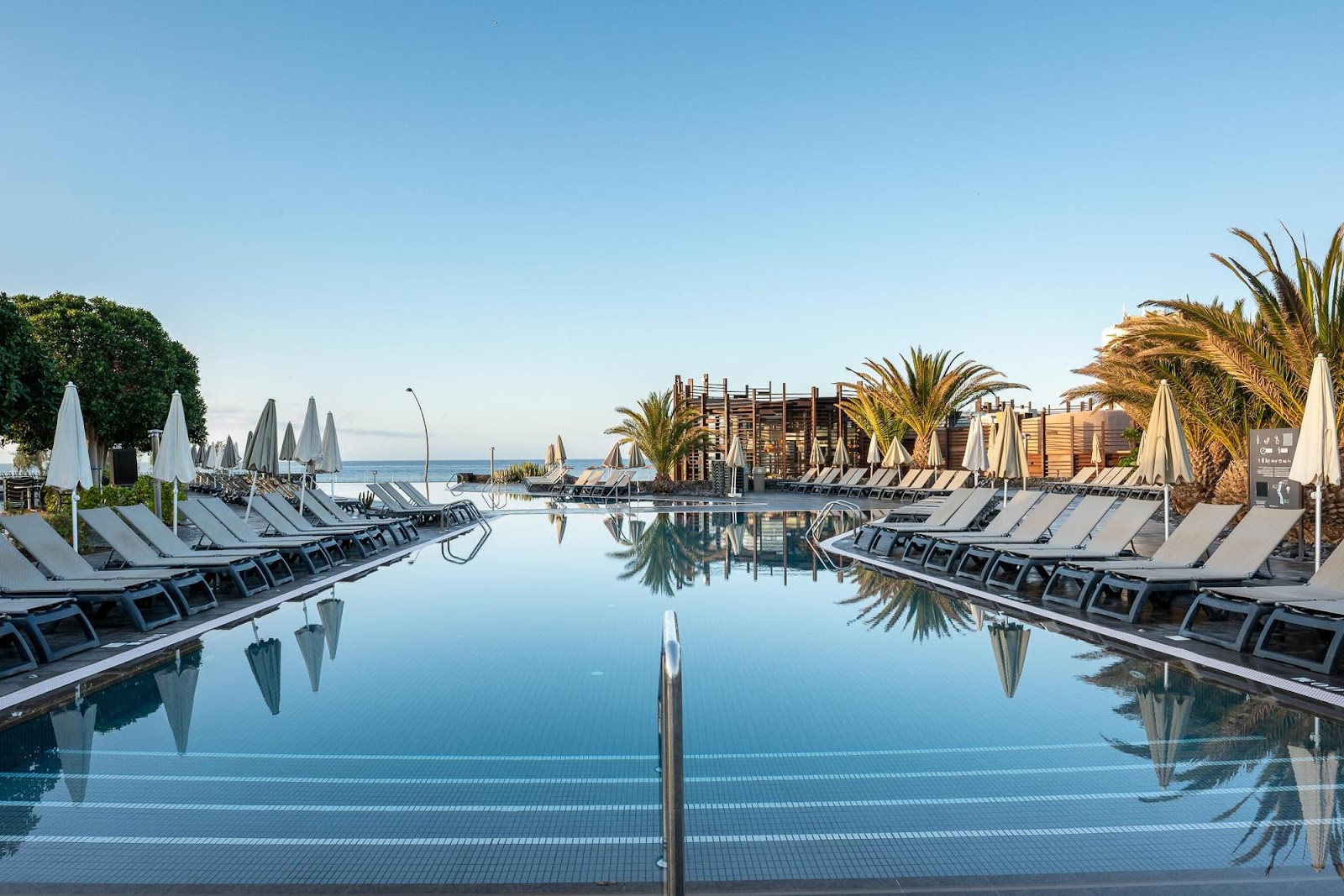 Resort pool lined with loungers and white umbrellas, palm trees and wooden structures visible beyond, ocean in background.