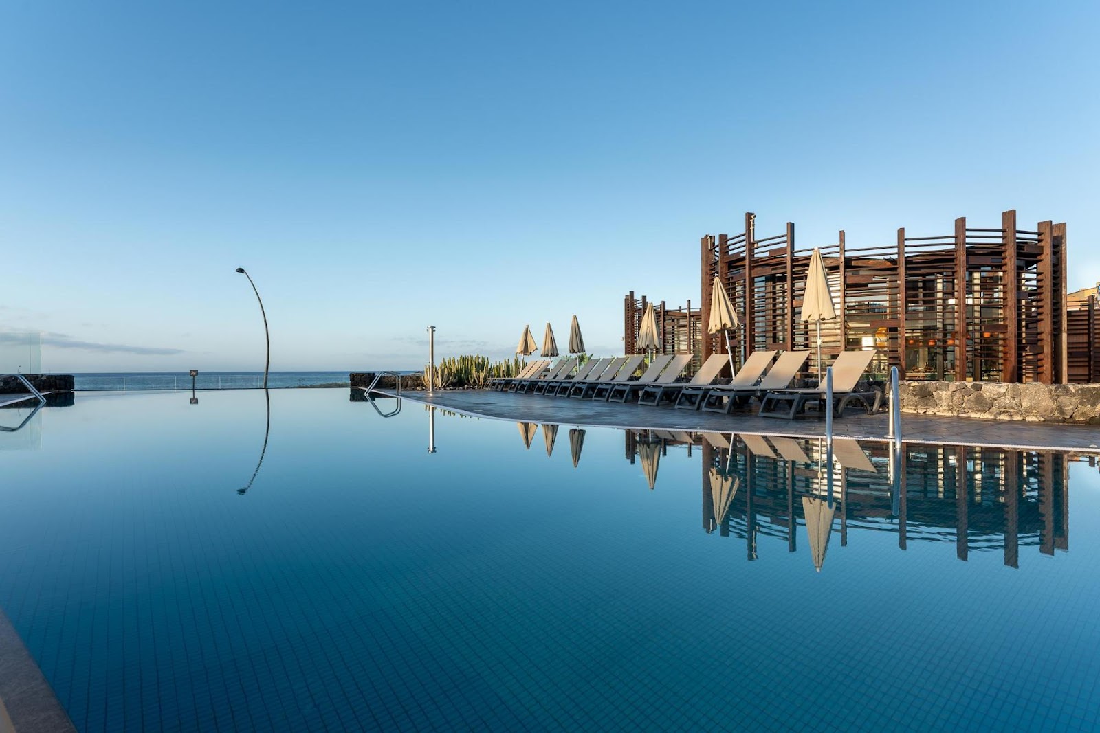 Wooden pergola structures with beige umbrellas and lounge chairs beside a calm pool overlooking the sea at Barceló Tenerife.