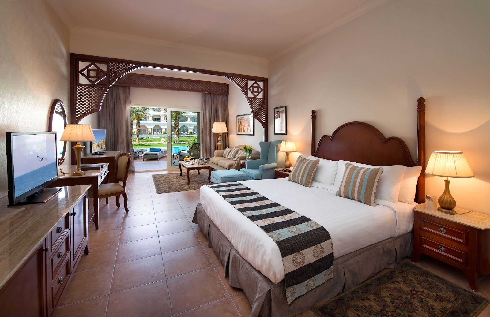 Resort suite with king bed, sitting area, and view of pool courtyard through open doors.