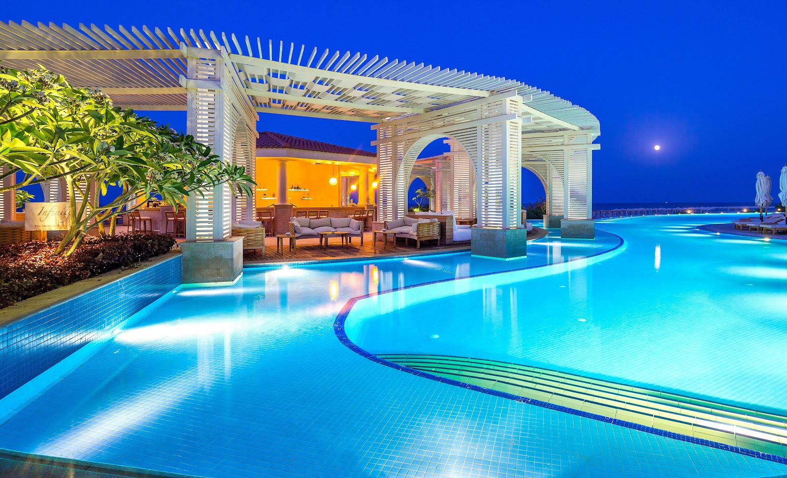 Illuminated curved pool with white pergolas and lounge seating at Baron Palace Sahl Hasheesh at dusk, overlooking the sea.