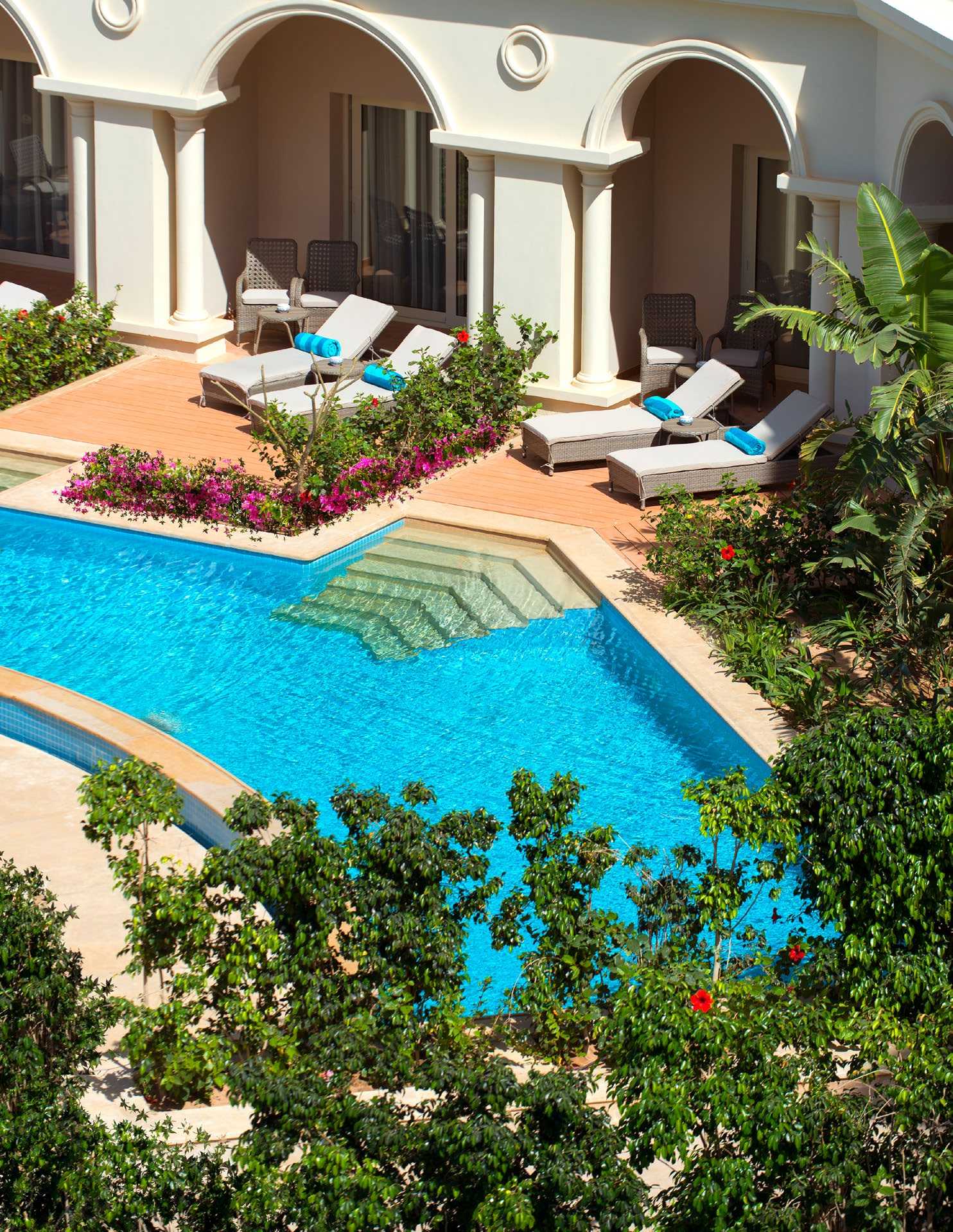 Blue pool with loungers and white umbrellas on terrace beside arched colonial-style building with magenta flowers.