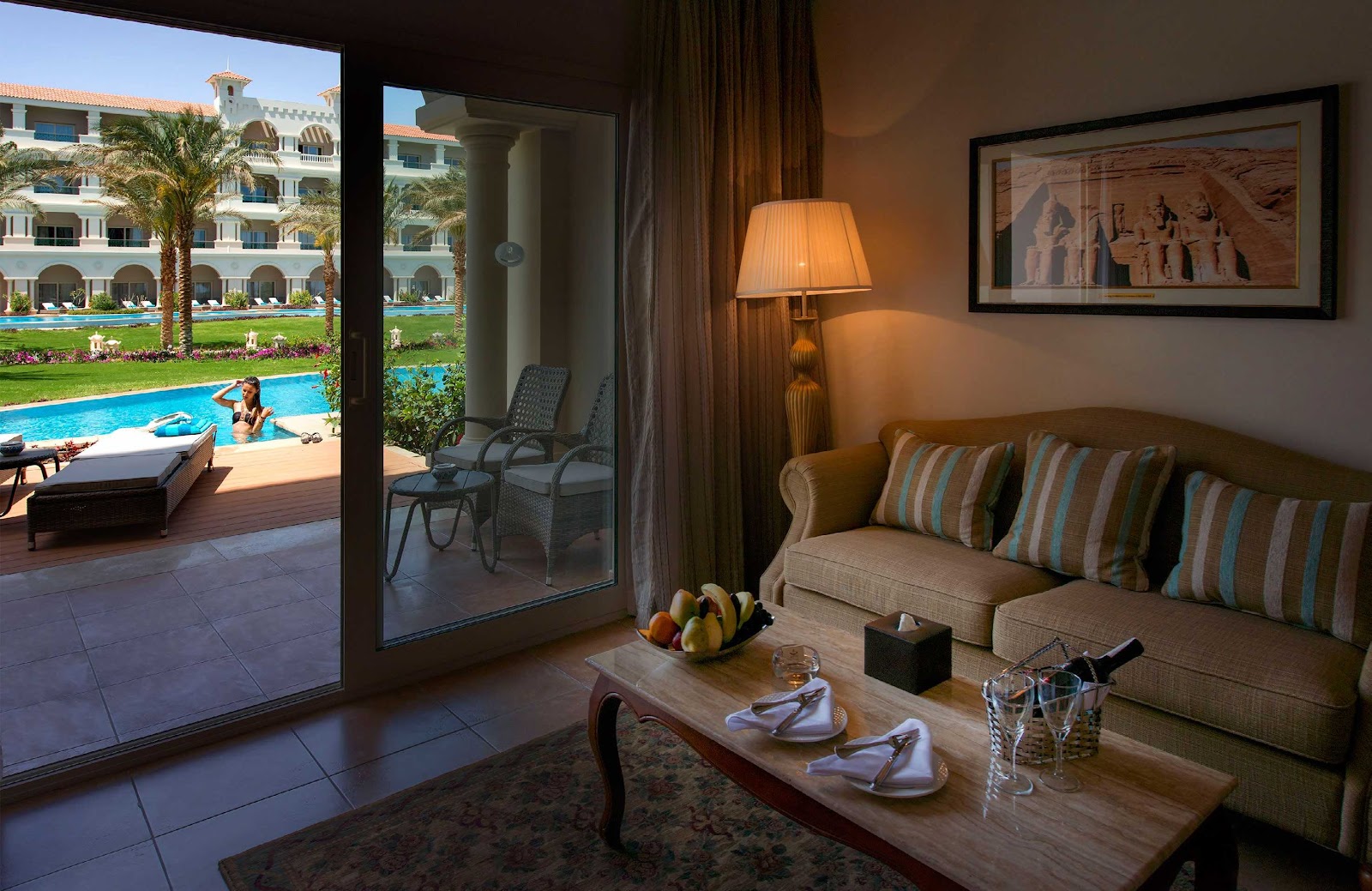 Curved beige seating surrounds a lit fire pit at dusk, overlooking a lagoon and palm trees at Baron Palace Sahl Hasheesh.