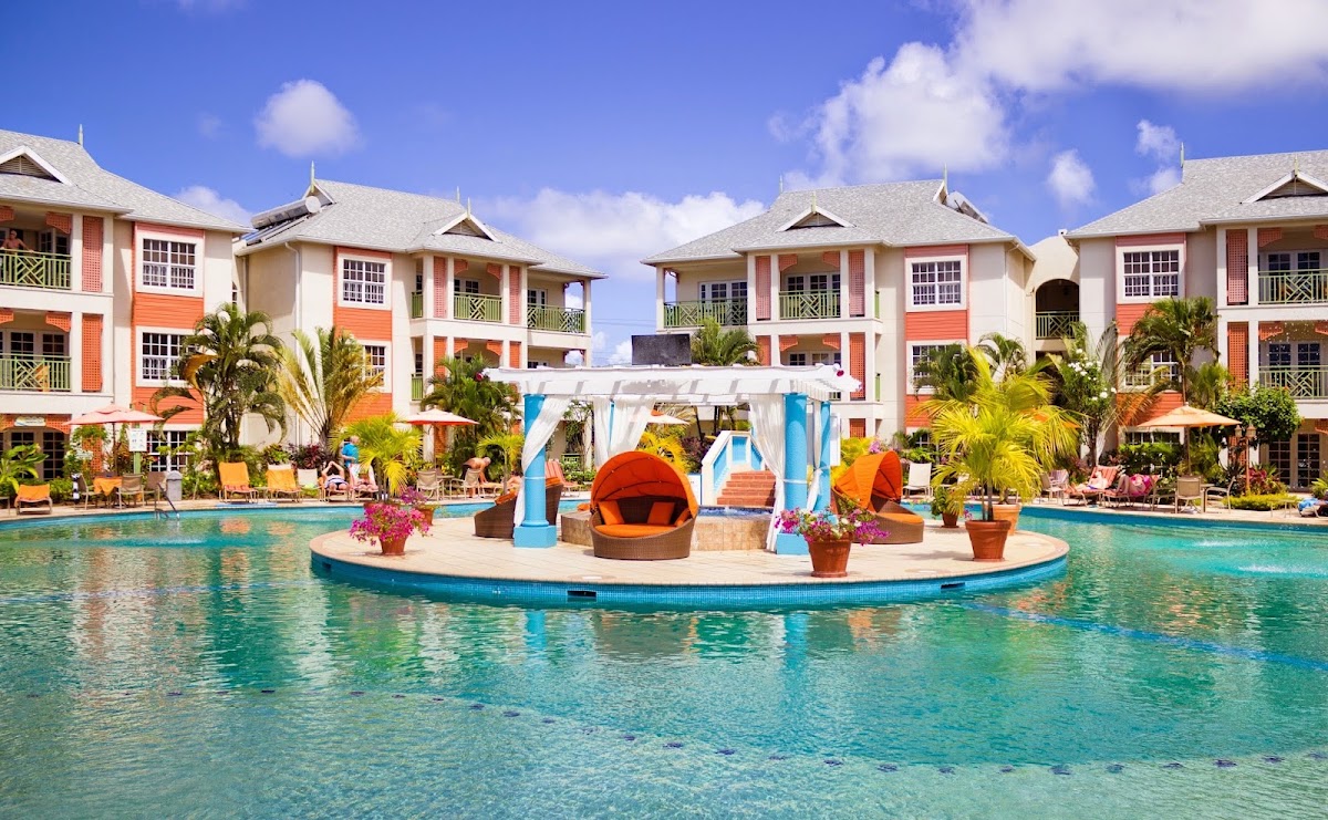 Colorful colonial-style resort buildings with orange and cream facades frame a lagoon pool with an island bar featuring orange pod seating.