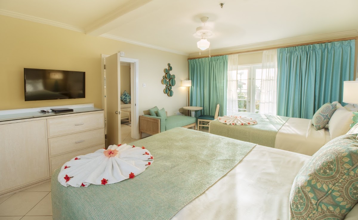 Resort room with two queen beds decorated with rose petals, turquoise curtains, wall-mounted TV, and balcony access.